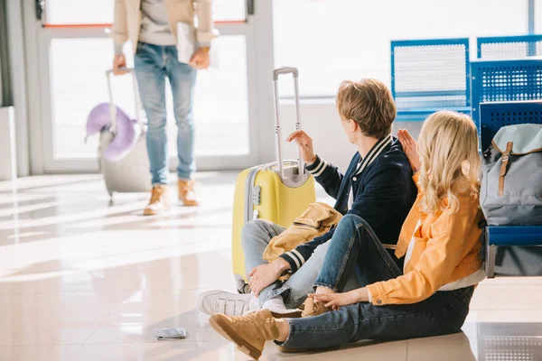 Junge Reisende, die auf dem Boden sitzen und Freunde anschauen, die am Flughafen hinter ihnen stehen — Stockfoto