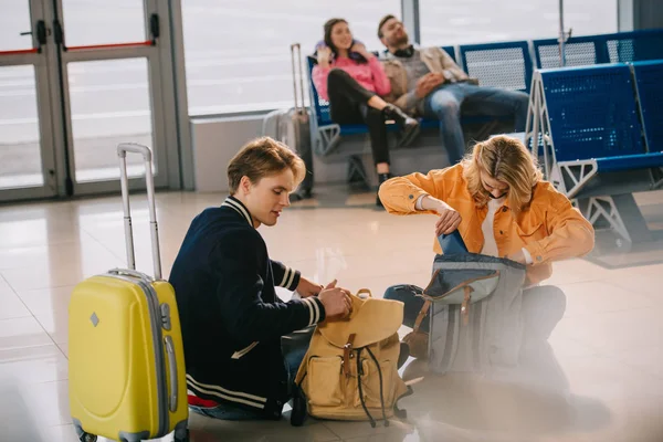 Jóvenes que miran en las mochilas mientras están sentados en el suelo y esperando el vuelo en el aeropuerto - foto de stock