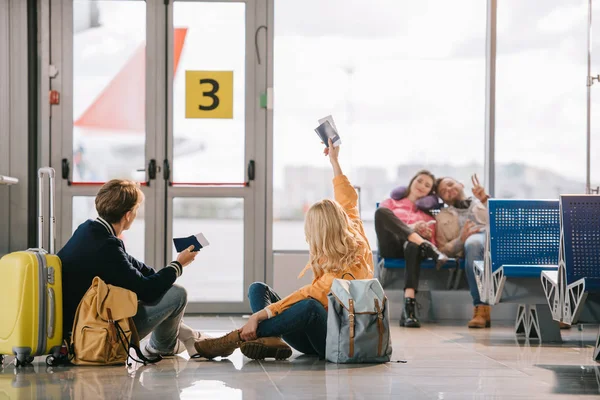 Jóvenes viajeros con pasaportes y tarjetas de embarque saludándose en la terminal del aeropuerto - foto de stock