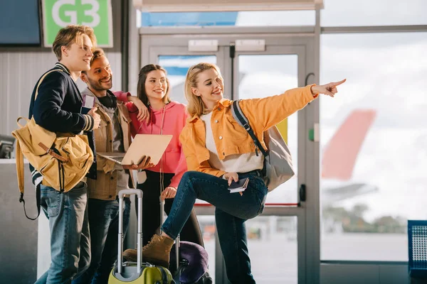 Jóvenes amigos sonrientes usando el ordenador portátil y apuntando hacia fuera mientras espera el vuelo en el aeropuerto - foto de stock