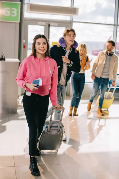 Mujer joven sonriente sosteniendo maleta, pasaporte y tarjeta de embarque mientras viaja con amigos - foto de stock