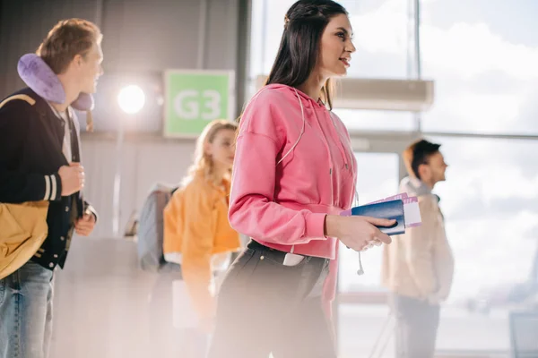 Vista de ángulo bajo de la chica sonriente sosteniendo pasaporte con tarjeta de embarque mientras va con amigos en la terminal del aeropuerto - foto de stock