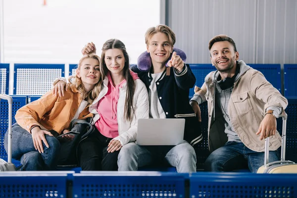 Happy young friends using laptop and smiling at camera while waiting for flight in airport terminal — Stock Photo