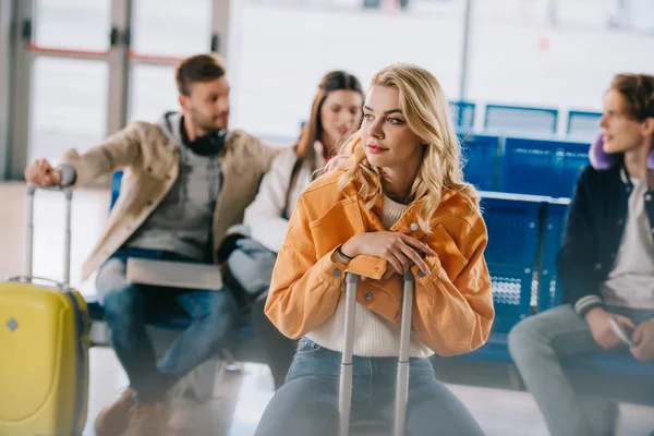 Chica sentada en la maleta y mirando hacia otro lado mientras espera con amigos en el aeropuerto — Stock Photo