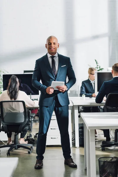 Young african american businessman using digital tablet and smiling at camera, colleagues working behind in office — Stock Photo