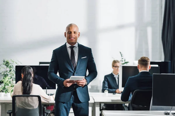 Young african american businessman holding digital tablet and smiling at camera, colleagues working behind in office — Stock Photo