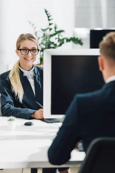 Cropped shot of businessman sitting at workplace and young businesswoman in eyeglasses smiling at camera — Stock Photo