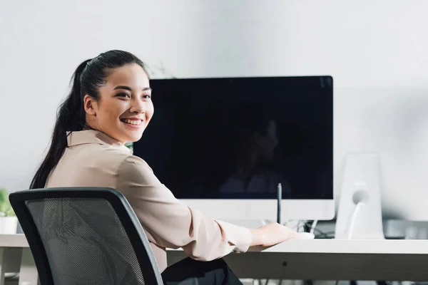 Beautiful smiling young businesswoman sitting at workplace and looking away in office — Stock Photo