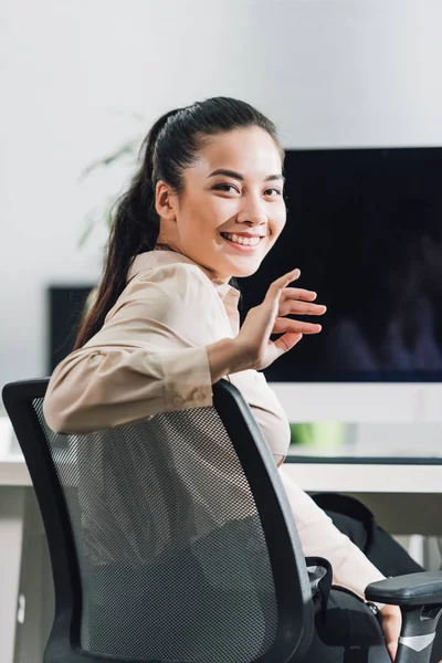 Красивая молодая деловая женщина машет рукой и улыбается в камеру во время работы в офисе — стоковое фото
