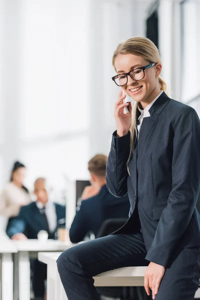 Lächelnde junge Geschäftsfrau mit Brille, die per Smartphone spricht und im Büro nach unten schaut — Stockfoto