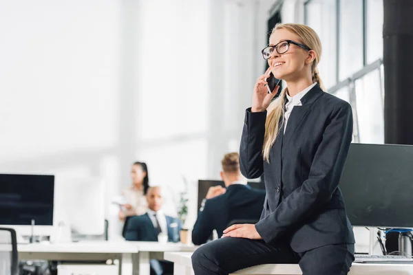 Sonriente joven mujer de negocios en gafas hablando por teléfono inteligente y mirando hacia otro lado en la oficina - foto de stock