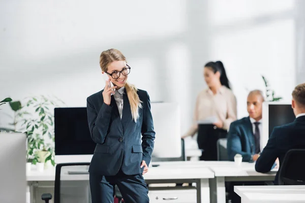 Lächelnde junge Geschäftsfrau mit Brille, die mit dem Smartphone spricht und in die Kamera schaut, während sie mit Kollegen im Großraumbüro arbeitet — Stockfoto