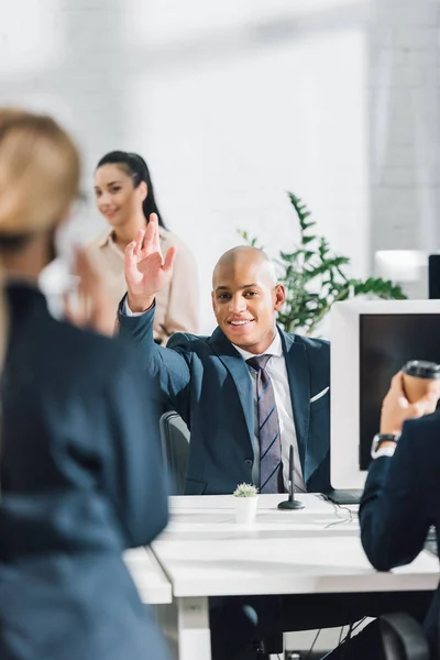 Joven hombre de negocios afroamericano saludando de la mano y sonriendo a la cámara mientras trabaja con colegas en la oficina de espacio abierto - foto de stock