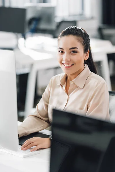 Beautiful young businesswoman using desktop computer and smiling at camera in office — Stock Photo