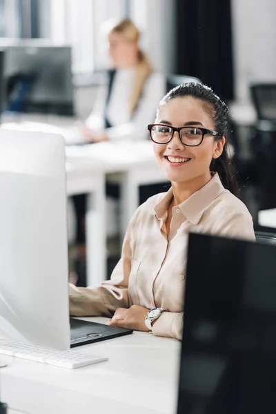 Attractive young businesswoman using desktop computer and smiling at camera in office — Stock Photo