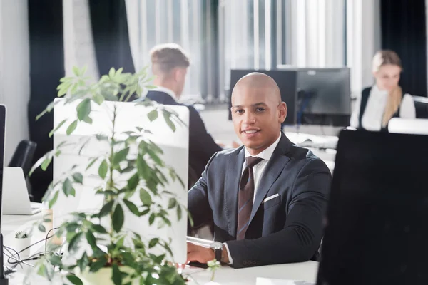 Guapo joven afroamericano hombre de negocios sonriendo a la cámara mientras trabaja en espacio abierto oficina - foto de stock