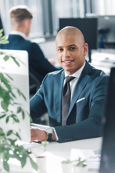 Handsome young african american businessman sitting at workplace and smiling at camera — Stock Photo