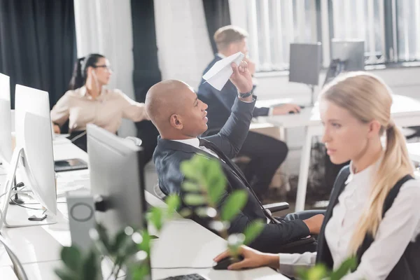 Giovane uomo d'affari afroamericano che gioca con l'aereo di carta mentre i colleghi lavorano nello spazio ufficio — Stock Photo