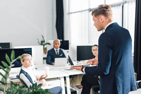 Serious young businessman using laptop and looking at colleagues in office — Stock Photo