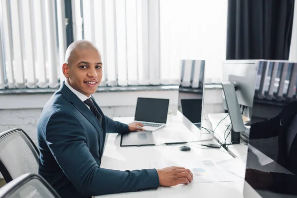 Bonito jovem Africano americano empresário sentado no local de trabalho e sorrindo para a câmera no escritório — Fotografia de Stock