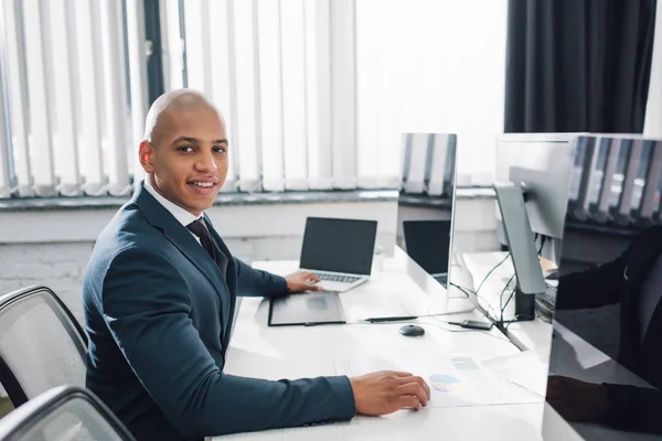 Guapo joven afroamericano hombre de negocios sentado en el lugar de trabajo y sonriendo a la cámara en la oficina - foto de stock