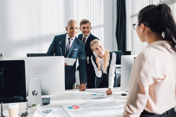 Sonriendo jóvenes compañeros de trabajo utilizando dispositivos digitales y mirando a una colega en la oficina - foto de stock