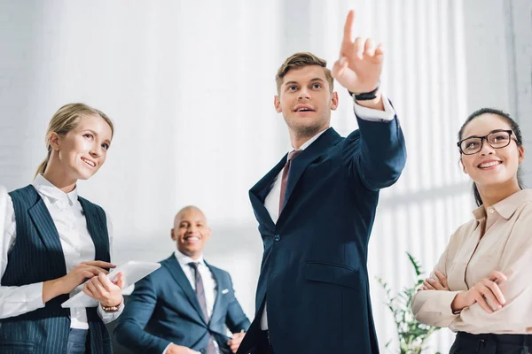 Smiling coworkers looking at young businessman pointing up with finger in office — Stock Photo