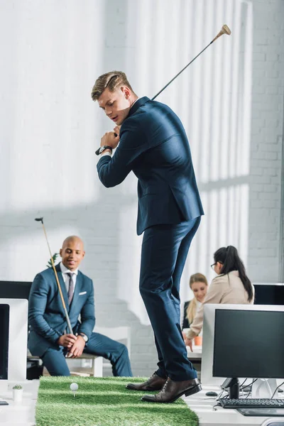 Jeunes hommes d'affaires multiethniques en tenue formelle jouant au golf au bureau — Photo de stock