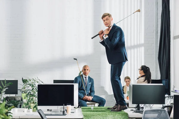 Jóvenes hombres de negocios multirraciales jugando al golf en una oficina de espacio abierto - foto de stock