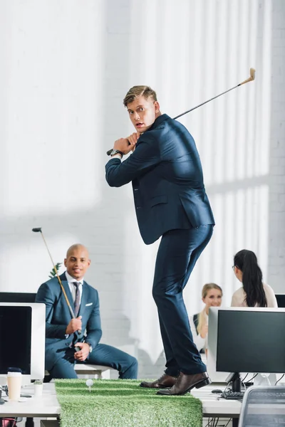 Jungunternehmer spielt Golf und Mitarbeiter sitzen im Büro hinten — Stockfoto