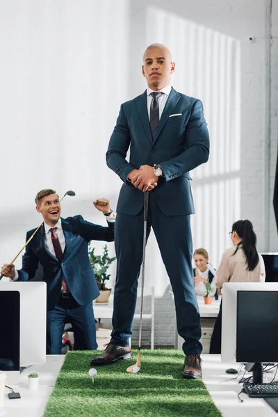 Confident young african american businessman looking at camera while playing golf with colleague in office — Stock Photo