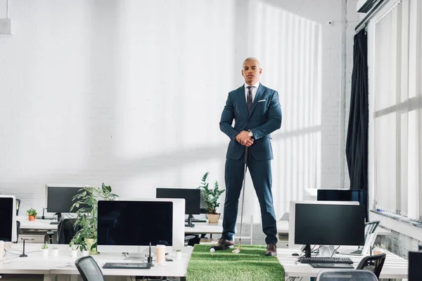 Confident young african american businessman standing on table and looking at camera while playing golf in office — Stock Photo