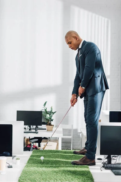 Vista lateral del joven hombre de negocios afroamericano jugando al golf en la oficina - foto de stock