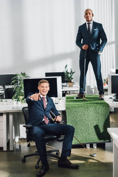 Glückliche junge multiethnische Geschäftsleute lächeln in die Kamera, während sie im Büro Golf spielen — Stockfoto