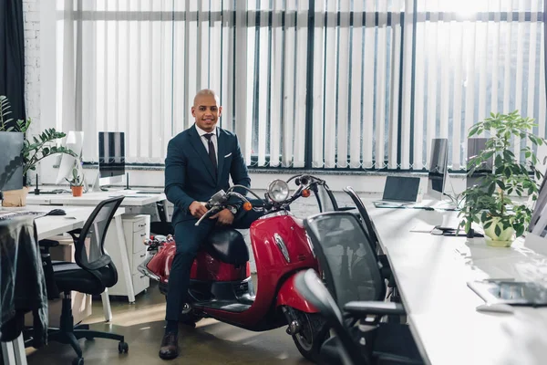 Feliz joven afroamericano hombre de negocios sentado en scooter rojo y sonriendo a la cámara en la oficina - foto de stock