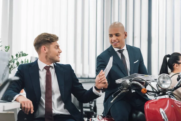 Sonriente joven hombre de negocios afroamericano sentado en scooter rojo y mirando a un colega masculino en la oficina - foto de stock
