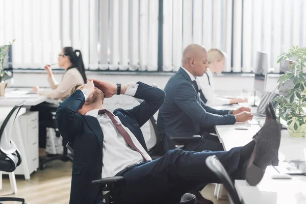 Young businessman rubbing eyes while sitting with legs on table at workplace — Stock Photo