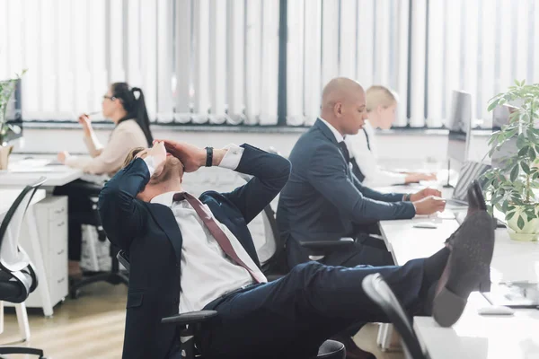 Joven hombre de negocios frotando los ojos mientras está sentado con las piernas en la mesa en el lugar de trabajo - foto de stock