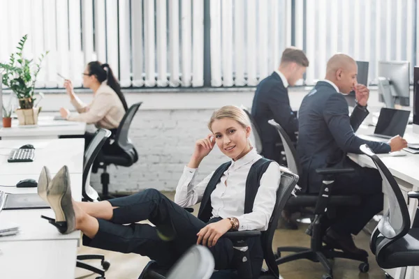 Hermosa joven empresaria sonriendo a la cámara mientras sus colegas trabajan detrás en la oficina de espacio abierto - foto de stock