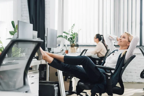 Улыбающаяся молодая деловая женщина отдыхает с ногами на столе в офисе — стоковое фото