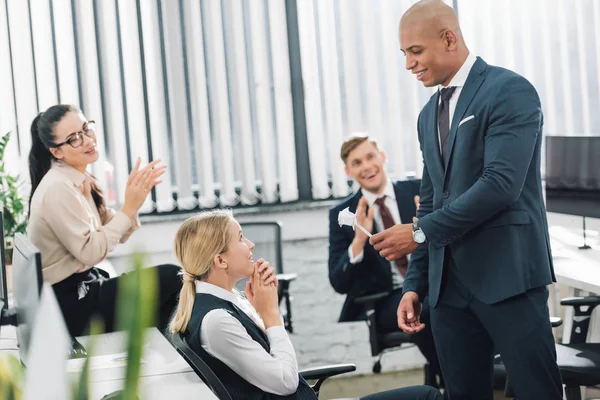 Beau jeune homme d'affaires afro-américain présentant fleur d'origami à femme d'affaires heureuse au bureau — Photo de stock