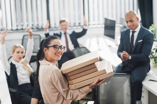 Junge Geschäftsfrau hält Pizzakartons in der Hand und lächelt in die Kamera, während Mitarbeiter im Großraumbüro hinter ihr sitzen — Stockfoto