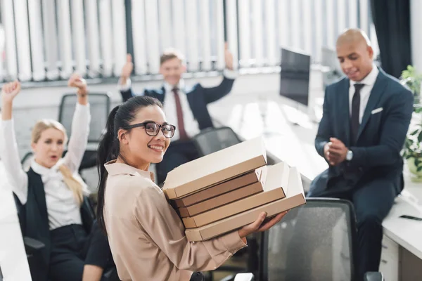 Joven empresaria sosteniendo cajas de pizza y sonriendo a la cámara mientras sus compañeros de trabajo se sientan detrás en una oficina de espacio abierto - foto de stock