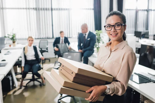 Joven empresaria sosteniendo cajas de pizza y sonriendo a la cámara mientras sus colegas se sientan detrás en la oficina - foto de stock