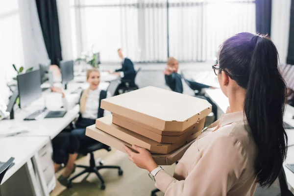 Mujer joven sosteniendo cajas de pizza y mirando a sus compañeros de trabajo en la oficina - foto de stock