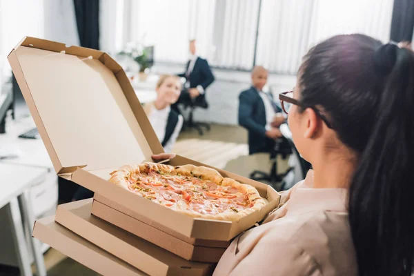 Junge Frau hält Pizzakartons in der Hand und schaut Kollegen im Großraumbüro an — Stockfoto