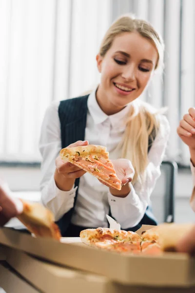 Sonriente joven empresaria comiendo pizza con colegas en la oficina - foto de stock