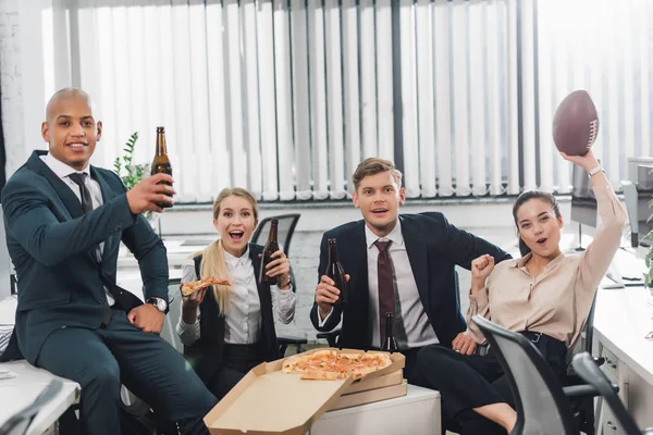 Happy young colleagues holding bottles of beer and smiling at camera in open space office — Stock Photo