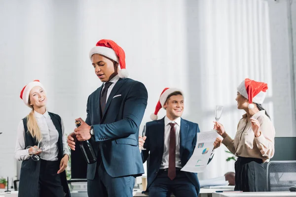 Junger afrikanisch-amerikanischer Geschäftsmann mit Weihnachtsmannhut öffnet Flasche Wein, während er mit Mitarbeitern im Büro das neue Jahr feiert — Stockfoto