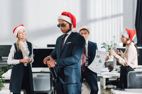 Young african american businessman in santa hat and sunglasses opening bottle of wine while celebrating christmas with coworkers in office — Stock Photo